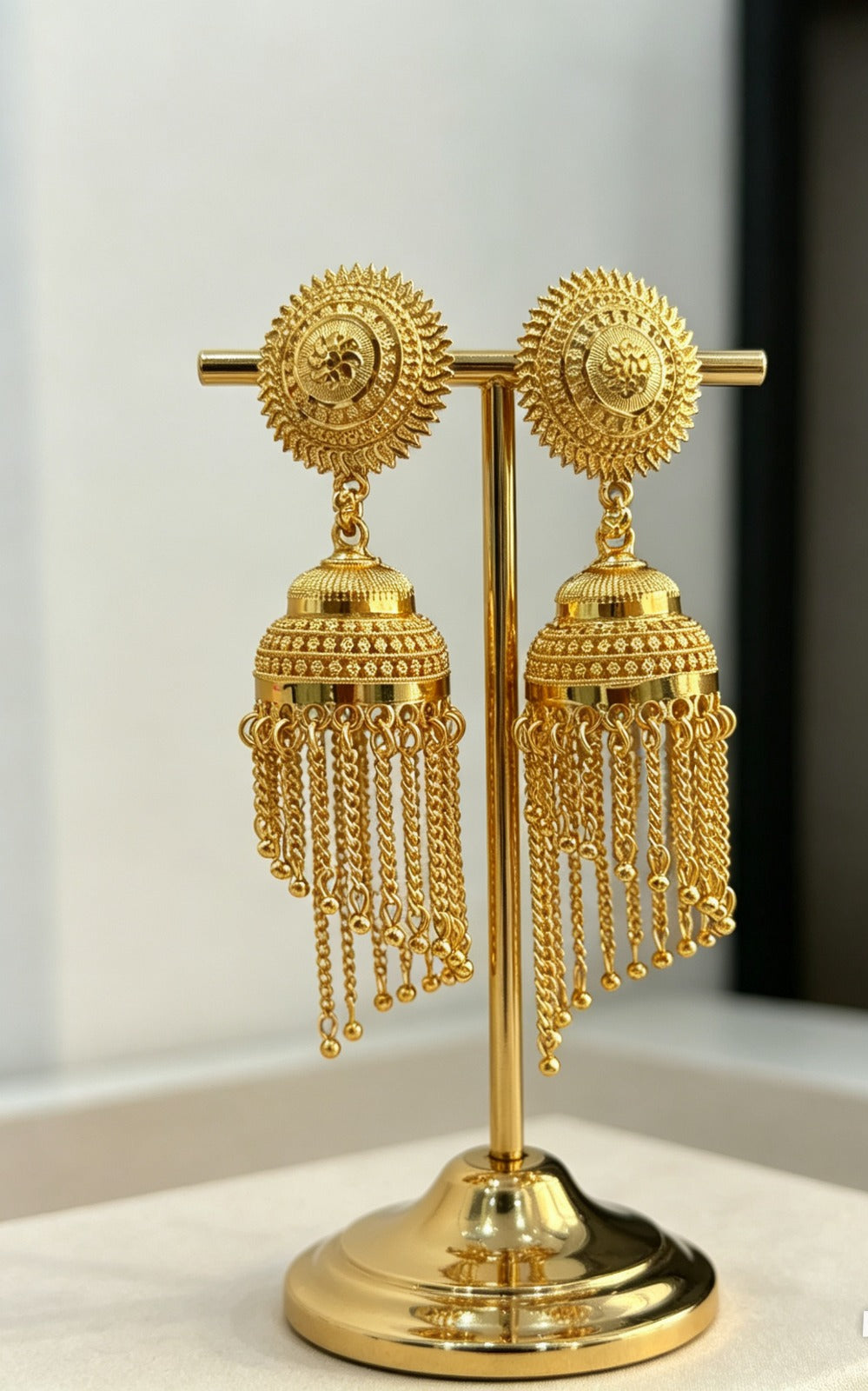 Pair of gold-plated jhumka earrings featuring intricate floral sunburst studs and hanging tiered chain tassels displayed on a premium gold jewelry stand in image.