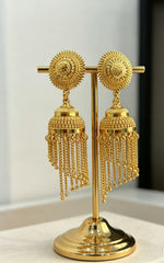 Pair of gold-plated jhumka earrings featuring intricate floral sunburst studs and hanging tiered chain tassels displayed on a premium gold jewelry stand in image.