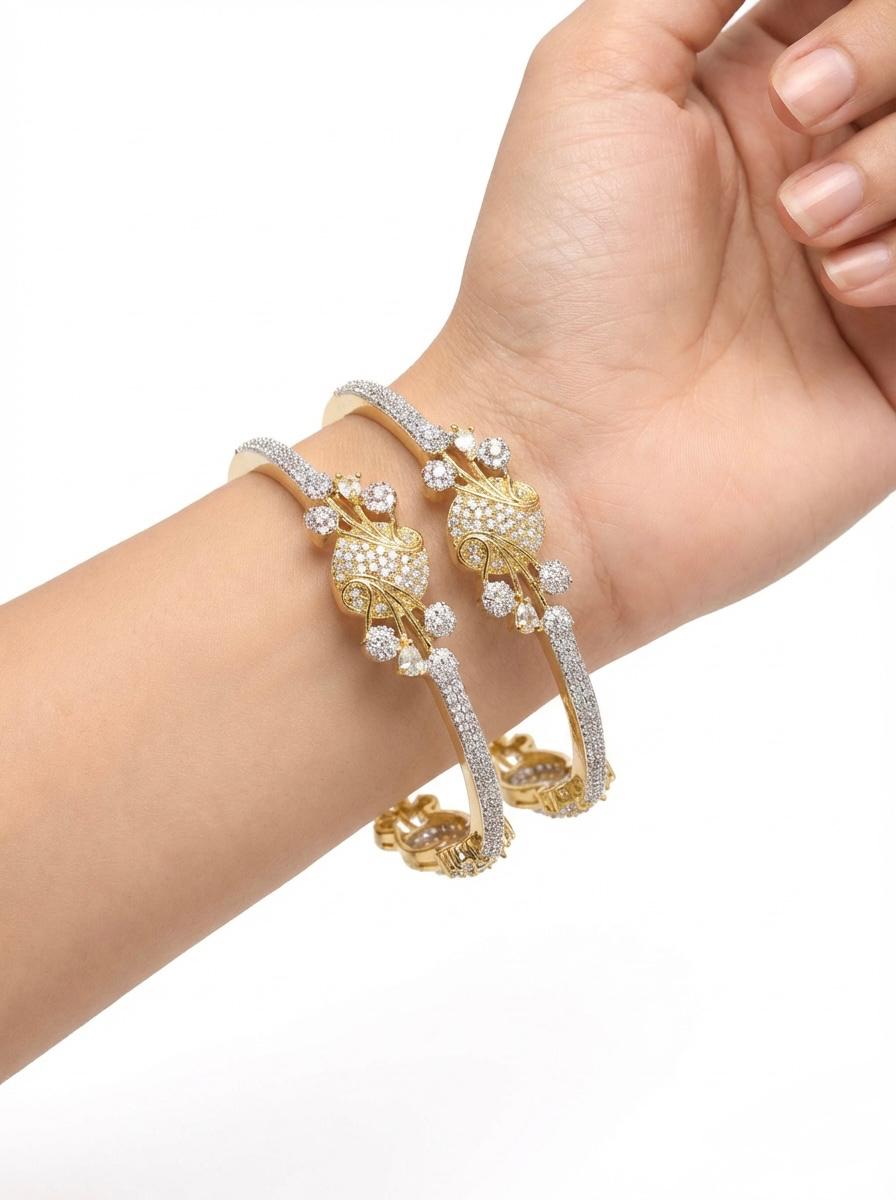 Close-up view of two gold-toned peacock motif bangles on a wrist, highlighting the brilliant cubic zirconia stonework and the secure side-opening mechanism.