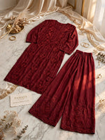 A two-piece women's outfit set in deep burgundy, featuring a long-sleeve top with floral embroidery and matching wide-leg pants, styled on a marble surface with gold rings, dried flowers, and sheer fabric.