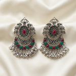 Regal Bloom silver-toned chandbali earrings with red and green stones, laid on a cream satin fabric background