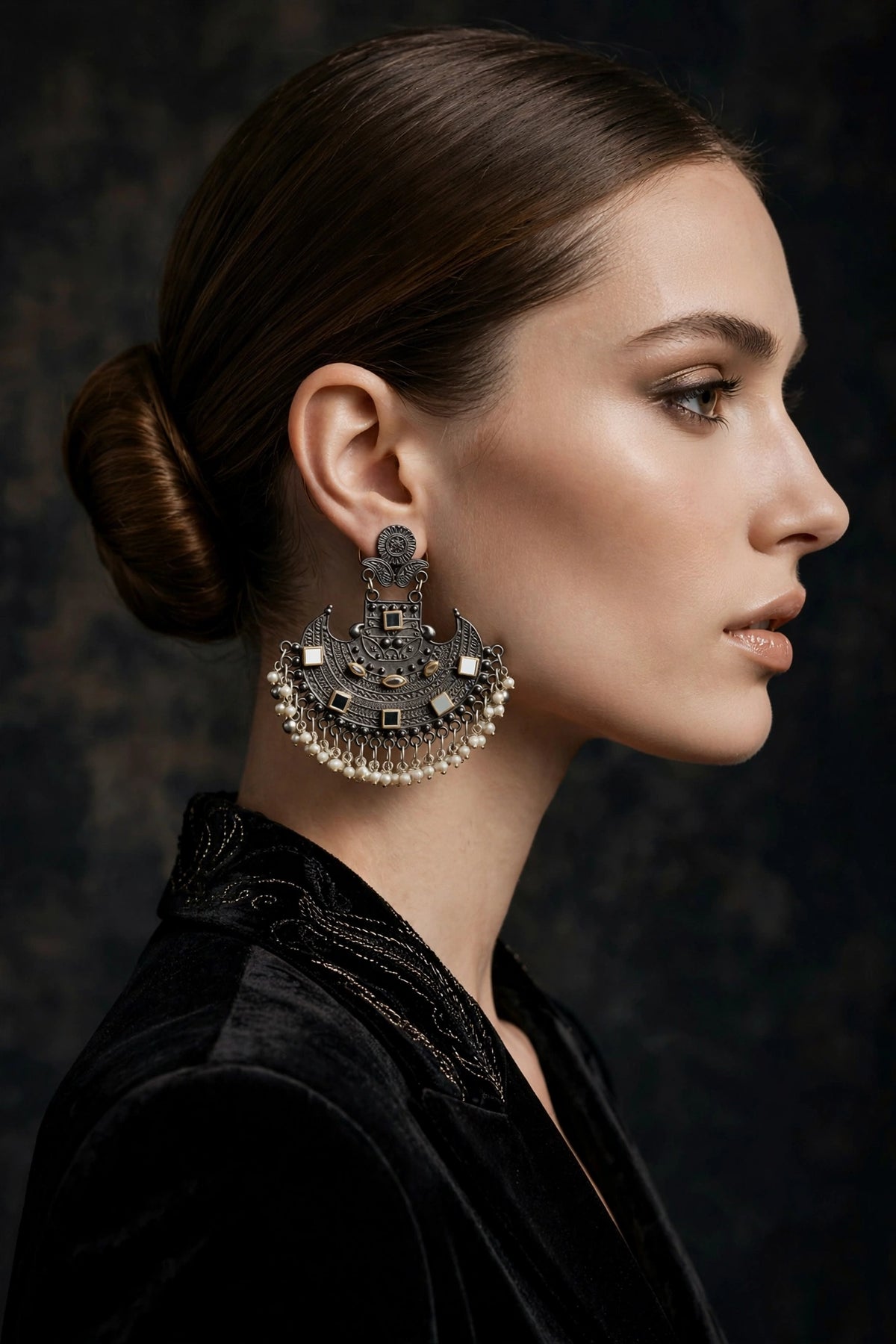 A close-up side profile of a woman with dark hair in a bun wearing a large, ornate silver-toned statement earring with intricate detailing and dangling pearl-like beads, set against a dark textured background.
