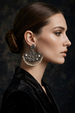 A close-up side profile of a woman with dark hair in a bun wearing a large, ornate silver-toned statement earring with intricate detailing and dangling pearl-like beads, set against a dark textured background.