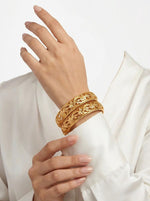 A professional photograph showing a model’s wrist wearing two intricate micro gold-plated copper floral vine bangles. The rich gold jewelry is contrasted by the crisp sleeve of a white silk blouse, highlighting the cutout pattern.