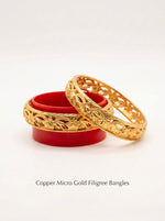 A pair of premium micro gold-plated copper bangles showcasing an openwork blossom and leaf pattern. One bangle rests atop a vibrant red circular display stand while the other leans against it, highlighting the high-polish finish and intricate cutout details against a clean white background.