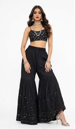 A model wearing a black cotton flared gharara featuring subtle tone-on-tone embroidery and delicate kaam dani detailing along the hem, styled with a matching patterned crop top and traditional jewelry against a white background.