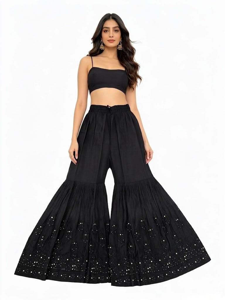Front view of a model in a black flared gharara, featuring subtle tone-on-tone embroidery and intricate Kaamdani work along the lower panel, paired with a simple black crop top against a white studio background.