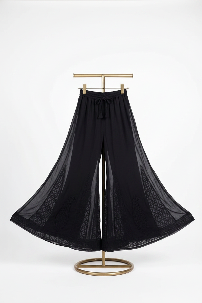 Black wide-leg pleated pants with a drawstring waist and sheer patterned side panels, displayed on a gold stand against a white background.