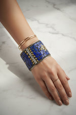 A close-up of a person's hand and wrist wearing a wide blue mosaic cuff bracelet with gold-toned metal details, alongside two slim gold bangles, set against a dark brown textured background with scattered lapis lazuli stones.