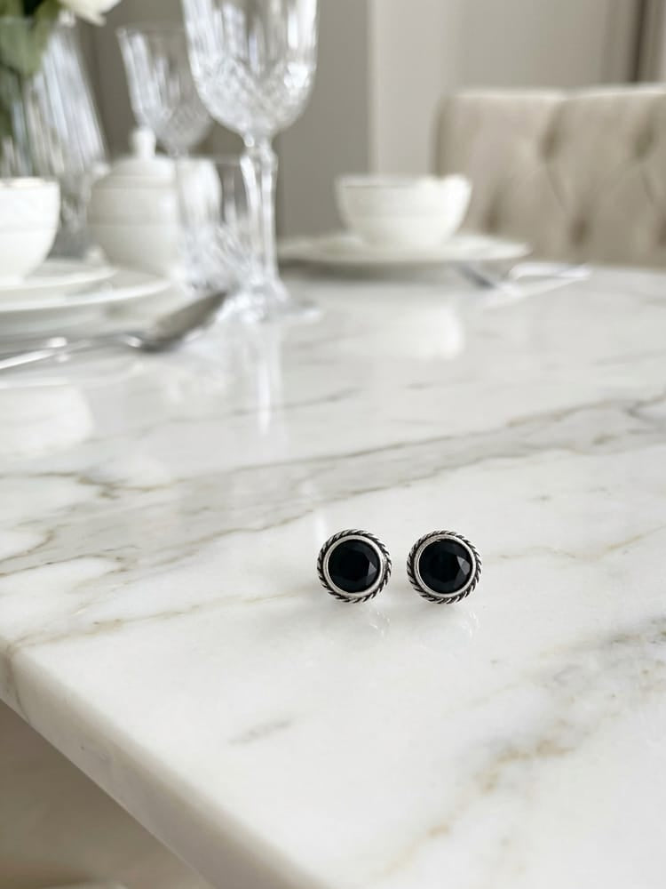 A pair of round black stud earrings with silver-toned textured borders, resting on a white marble dining table with a blurred background of a dining room setting.
