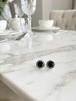 A pair of round black stud earrings with silver-toned textured borders, resting on a white marble dining table with a blurred background of a dining room setting.
