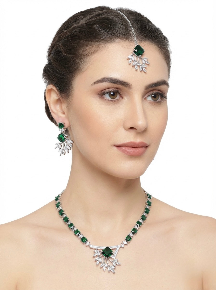 Model is wearing American diamond jewellery set including necklace, earrings and maang tikka with emerald green centre stone. 