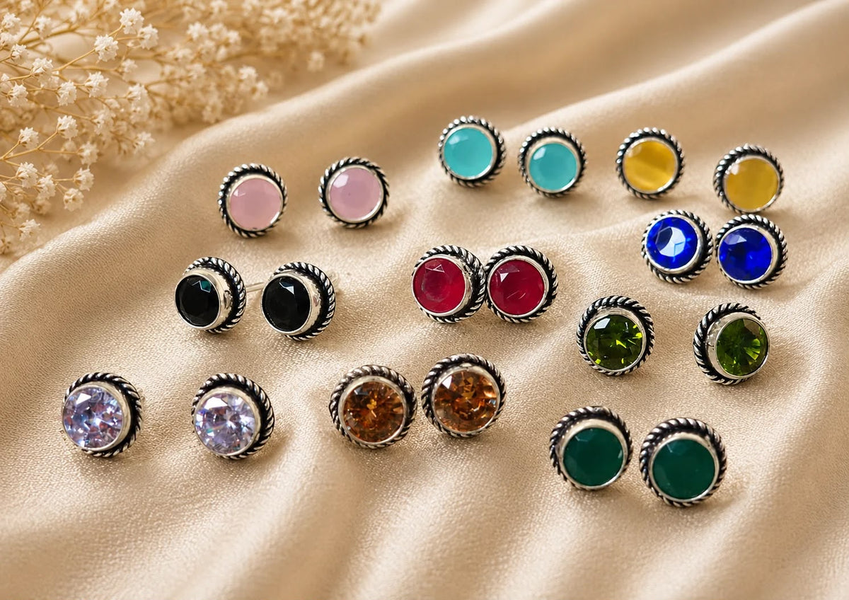 A collection of round bezel-set stud earrings with silver-toned twisted borders and various colored gemstones, arranged on draped beige satin fabric with dried flowers in the corner.