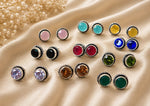 A collection of round bezel-set stud earrings with silver-toned twisted borders and various colored gemstones, arranged on draped beige satin fabric with dried flowers in the corner.