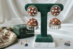 A pair of ornate silver-toned jhumka earrings decorated with colorful multicolor beads, displayed on a dark green velvet T-stand on a wooden table.