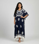 A navy blue Chikankari kaftan dress with intricate white thread embroidery and a V-neckline, standing by a bench with grass and flowers in the background.
