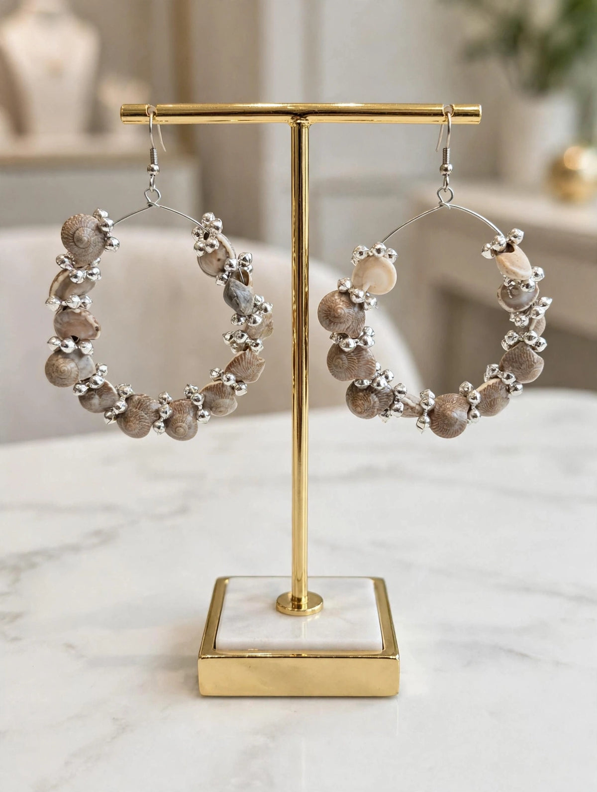 Neutral beige seashell hoop earrings with silver-toned beads displayed on gold and white marble stand, handcrafted boho design.