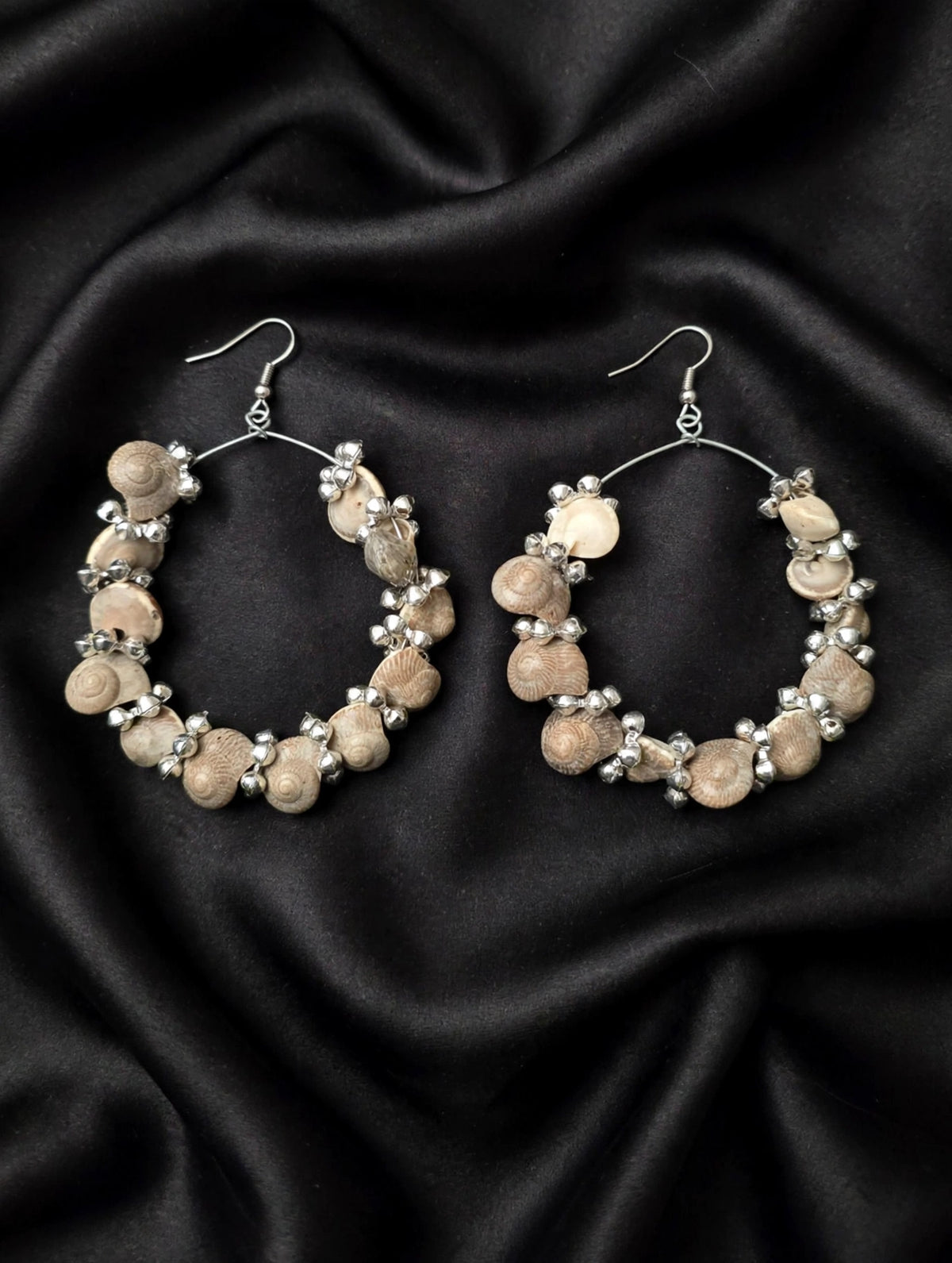 Pair of neutral beige seashell hoop earrings with silver-toned beads laid flat on luxurious black satin fabric, handcrafted boho design.