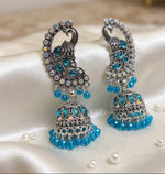 A pair of ornate silver-toned jhumka earrings embellished with blue and iridescent rhinestones, displayed on draped white satin fabric with scattered decorative pearls.