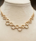 A close-up view of a luxury 18k gold plated honeycomb geometric necklace displayed on a soft, neutral stand, showcasing the elite craftsmanship and regal grandeur of the design.