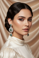 A close-up portrait of a woman wearing a pair of dramatic drop earrings with green stones and clear crystal-like settings, paired with a cream satin high-neck top against a draped beige fabric background.