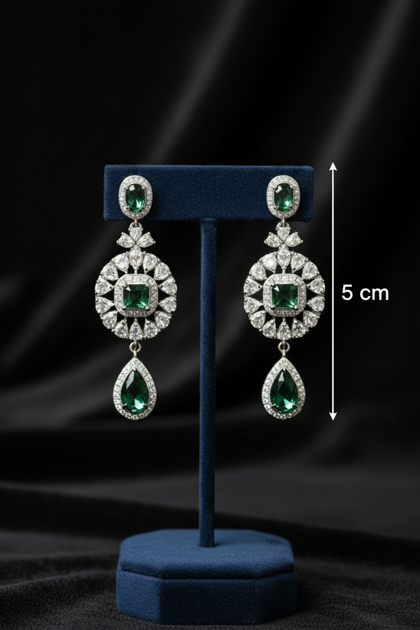A pair of silver-toned drop earrings with green gemstones and clear stones, displayed on a dark blue stand against a black satin background, with a 5 cm height marker shown on the right.