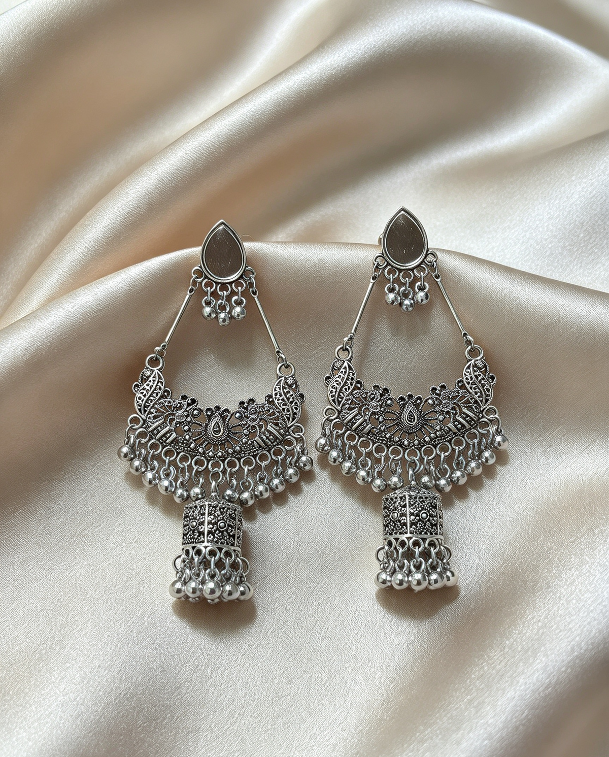 Symmetric pair of oxidized silver-tone chandbali jhumka earrings — identical size, intricate filigree peacock detailing, cascading ghungroo beads, lantern jhumka drop, displayed on soft cream satin surface.
