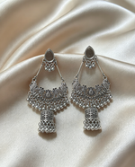 Symmetric pair of oxidized silver-tone chandbali jhumka earrings — identical size, intricate filigree peacock detailing, cascading ghungroo beads, lantern jhumka drop, displayed on soft cream satin surface.