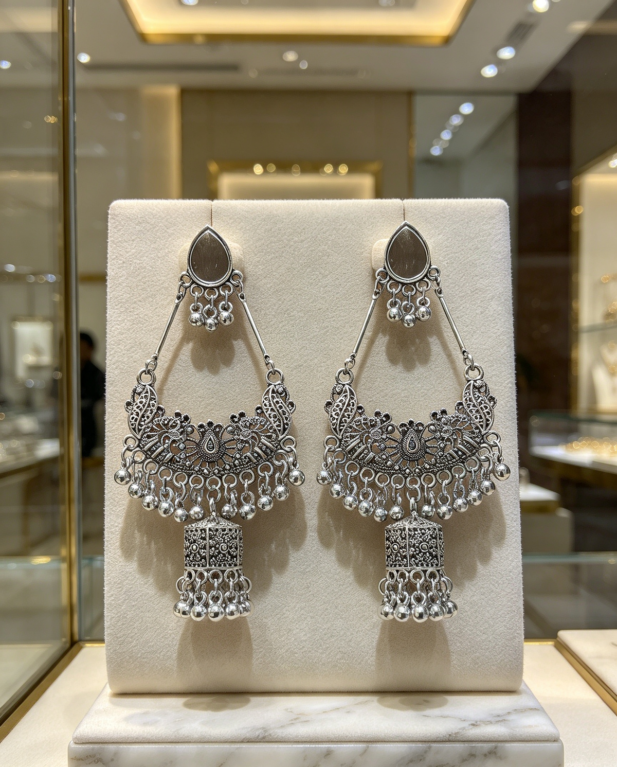 Symmetric pair of oxidized silver-tone chandbali jhumka earrings — identical size, intricate filigree, peacock detailing, dangling ghungroo beads, lantern drop, displayed on cream stand in premium jewelry boutique.