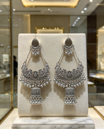 Symmetric pair of oxidized silver-tone chandbali jhumka earrings — identical size, intricate filigree, peacock detailing, dangling ghungroo beads, lantern drop, displayed on cream stand in premium jewelry boutique.