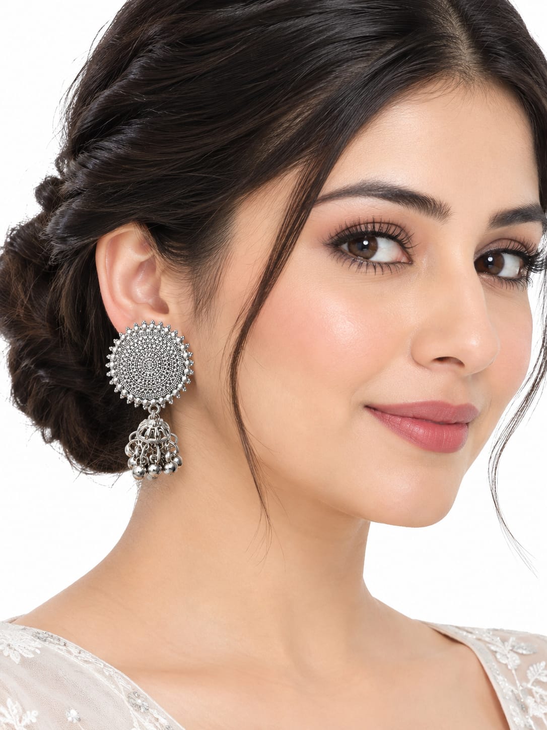 A close-up of a woman wearing a large silver-toned Indian jhumka earring with a detailed circular disc and dangling bell charm, set against a plain white background.