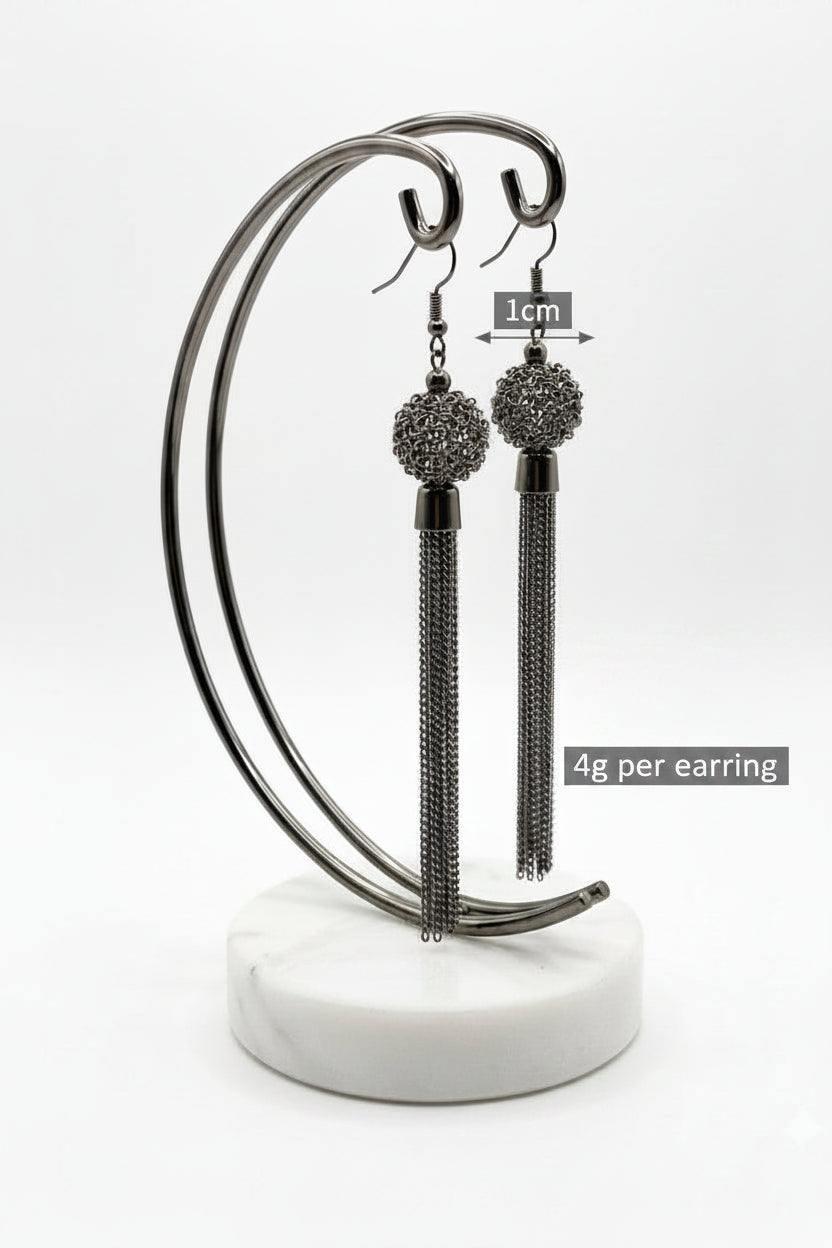 A professional product photograph of a pair of high-fashion dangle earrings displayed on a dark metallic curved stand with a white marble base. The gunmetal-finished earrings feature intricate, spherical filigree wire mesh balls from which long, cascading multi-strand fine chain tassels hang, set against a seamless white background.
