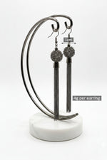 A professional product photograph of a pair of high-fashion dangle earrings displayed on a dark metallic curved stand with a white marble base. The gunmetal-finished earrings feature intricate, spherical filigree wire mesh balls from which long, cascading multi-strand fine chain tassels hang, set against a seamless white background.