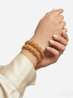 A lifestyle photograph of a woman's wrist wearing two micro gold-plated copper bangles featuring three intricate hollow filigree orbs and a textured band, styled with a cream silk blouse.