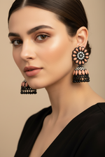 Model is wearing Pair of black brass jhumka earrings displayed on a stand, showcasing a soft peach floral stone stud and matching peach-accented jhumka base.