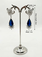 Pair of earrings with blue teardrop-shaped stones on a stand with measurements.