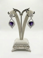 Pair of heart-shaped purple gemstone earrings on a decorative stand against a white background