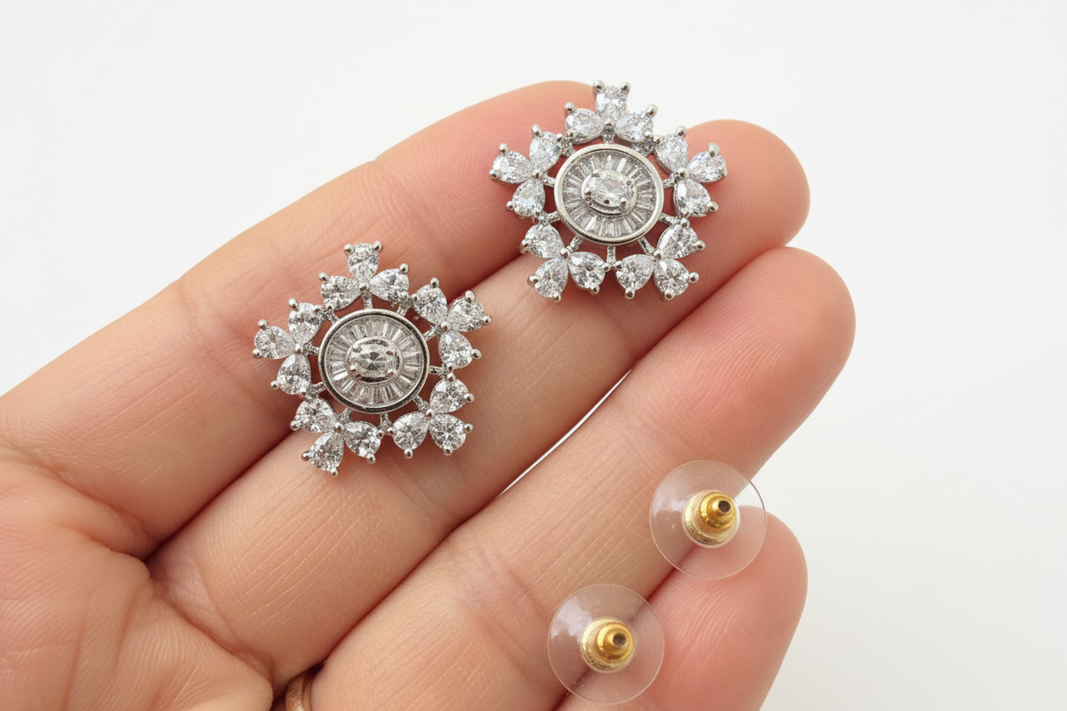 A pair of silver-toned diamond stud earrings with a sunburst floral design and a central round diamond, held on a person's fingers with two clear display backs visible below.