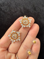 A close-up of a hand holding two gold-toned stud earrings with clear gemstone sunburst designs, with two additional earring backs visible on clear stands against a dark textured background.