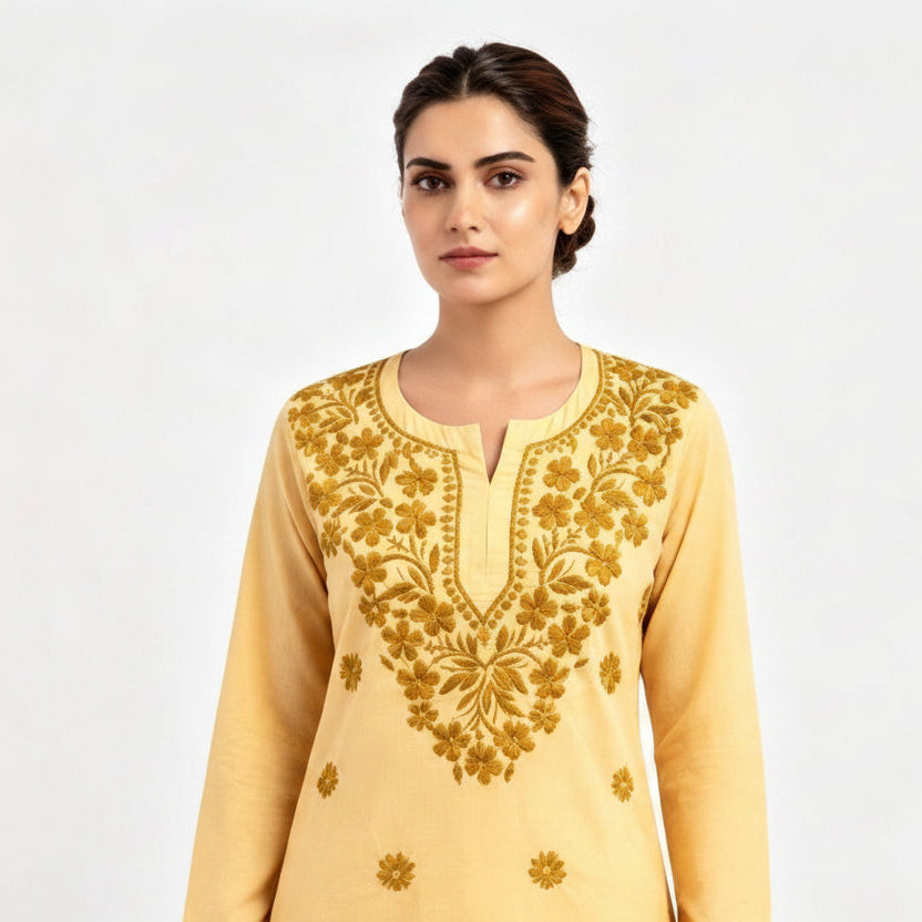 A woman modeling a pale yellow long-sleeve short kurti with intricate gold floral embroidery on the chest and scattered motifs on the lower front, paired with white trousers against a plain light background.