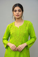 Front view of a woman wearing a vibrant parrot green cotton short kurti featuring intricate matching floral embroidery, a keyhole neckline, and a square hem line, styled with traditional earrings.