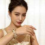 Model wearing Famaza Couture gold-tone open cuff bracelet with two lustrous white pearls and sparkling clear pavé crystals, ornate layered design, styled with champagne satin gown in luxury marble gold interior — elegant bridal and formal statement jewellry.