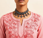 Close-up view of the neckline and chest area of a blush pink cotton short kurti featuring dense, intricate pink floral embroidery and a keyhole neckline, styled with an ornate silver choker necklace.