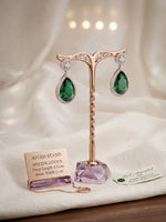 A posh brand jewelry display stand in rose gold featuring an amethyst base, showcasing a pair of emerald teardrop earrings with a 4.5 cm drop length.