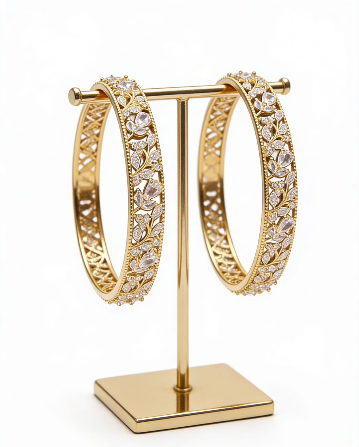 A pair of premium handcrafted gold-plated American Diamond bangles featuring an intricate leaf and vine motif, displayed on a minimalist gold T-stand.