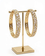 A pair of premium handcrafted gold-plated American Diamond bangles featuring an intricate leaf and vine motif, displayed on a minimalist gold T-stand.