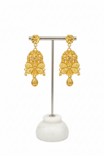 A pair of ornate gold-toned dangle earrings displayed on a silver metal stand with a white marble base against a plain white background.