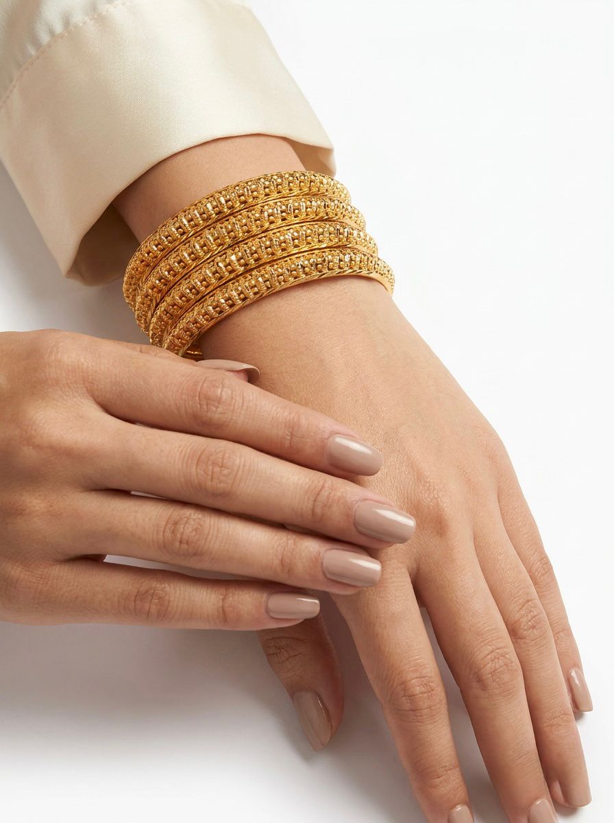 A lifestyle photograph of a model's hand wearing a set of four intricate, micro gold-plated textured bangles. The bangles feature a detailed lattice and beaded pattern, paired with a sophisticated white silk sleeve and neutral-toned manicure against a clean white background.