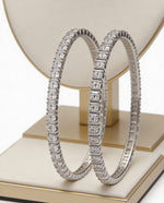 A pair of premium silver-plated American diamond bangles with a precision single-row square setting, elegantly displayed on a modern gold and cream luxury jewelry stand.