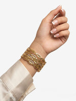 A lifestyle shot of a woman’s wrist adorned with four three-tone micro gold-plated bangles featuring an intricate floral lattice design in yellow, white, and pink gold finishes. She is wearing a cream-colored silk blouse against a soft white background.